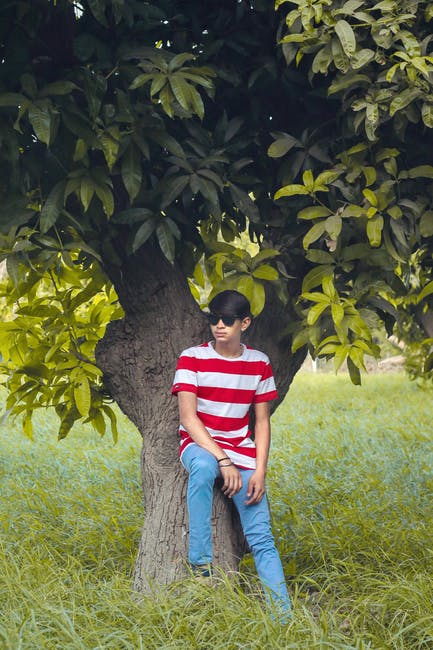 Man Wearing White和红色条纹船员衬衫-蓝色牛仔牛仔裤-黑色旅行者式太阳镜坐在树上