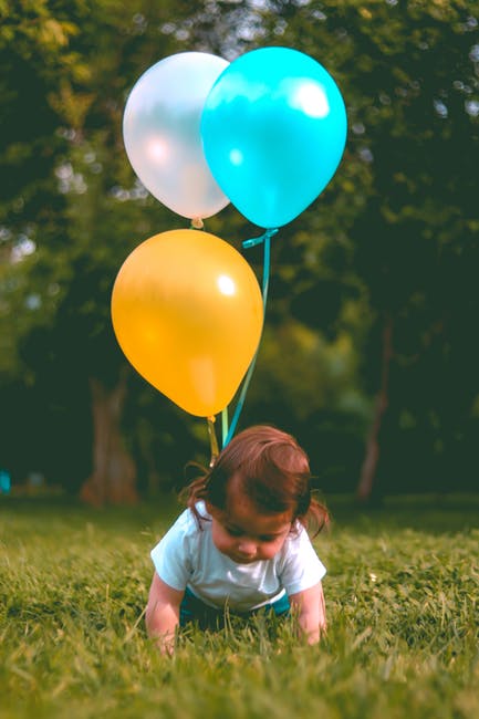 婴儿穿着白色T恤-在森林附近的绿草地上拿着三个黄色-蓝色和白色的气球