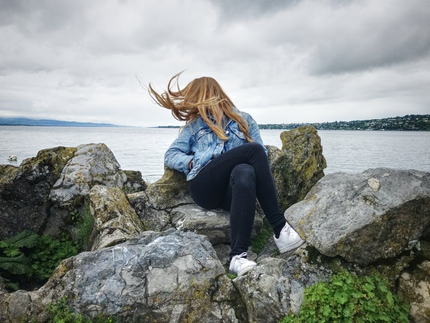 穿着蓝色牛仔裤的女式紧身上衣和黑色牛仔裤坐在灰色的岩石上观看湖景