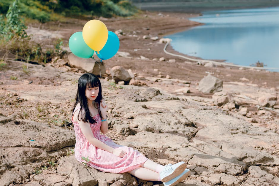 女人穿着粉色厚背带裙-坐在岩石地面上拿着黄色和蓝色的气球