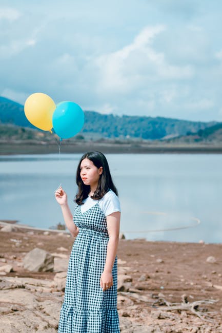 妇女穿着蓝白相间的格子裙-白天在平静的水体旁拿着蓝色和黄色的气球。
