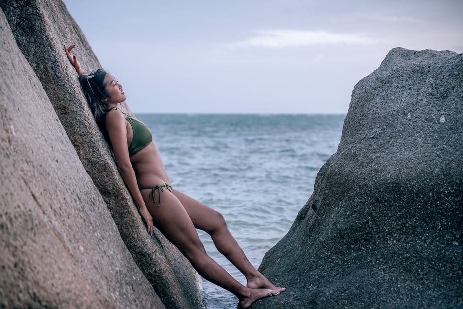 身穿绿色比基尼的女人倚在水体旁的岩石上