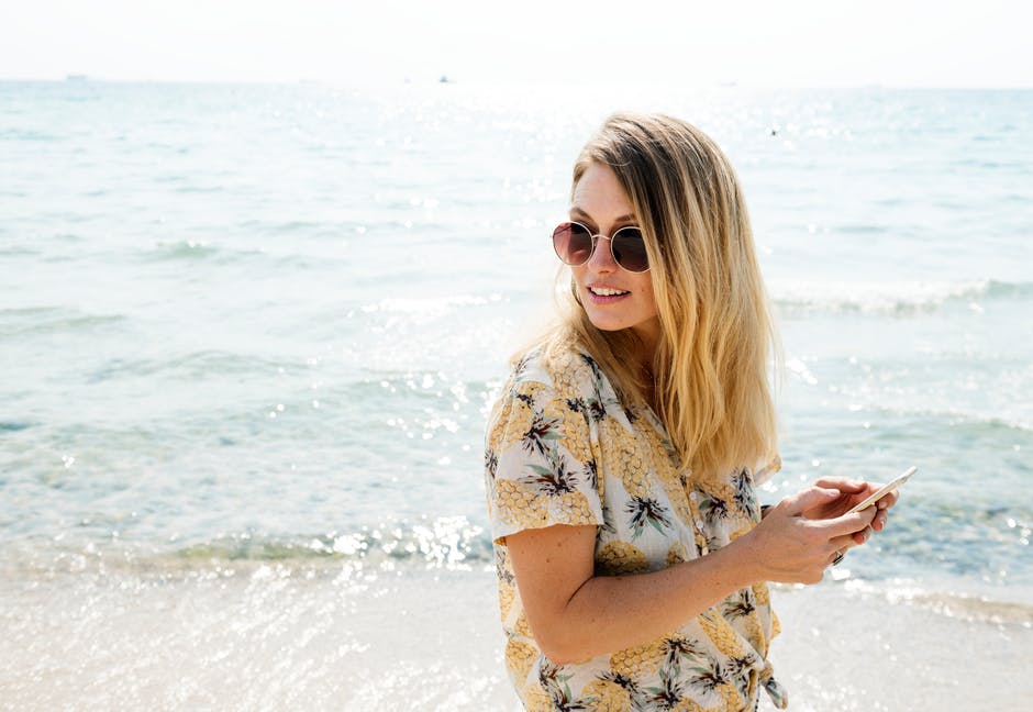 身穿黑色黄白相间钮扣衬衫的女子手持智能手机戴着太阳镜靠近水体
