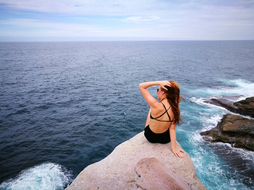 穿着黑色泳装的女人坐在灰色的石质悬崖面向水面